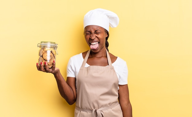 Black afro chef woman with cheerful, carefree, rebellious attitude, joking and sticking tongue out, having fun. cookies concept