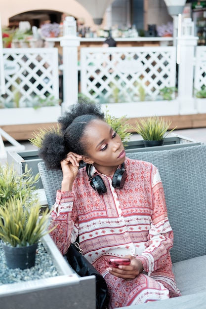 Black afro girl in ethnic dress with headphones on the neck sitting in open air cafe, holding smartphone and thinking about something