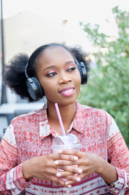 Black afro girl in ethnic dress with headphones sitting in open air cafe, listening to music and drinking milk cocktail