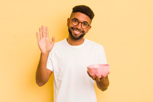 Black afro man smiling happily waving hand welcoming and greeting you empty bowl concept