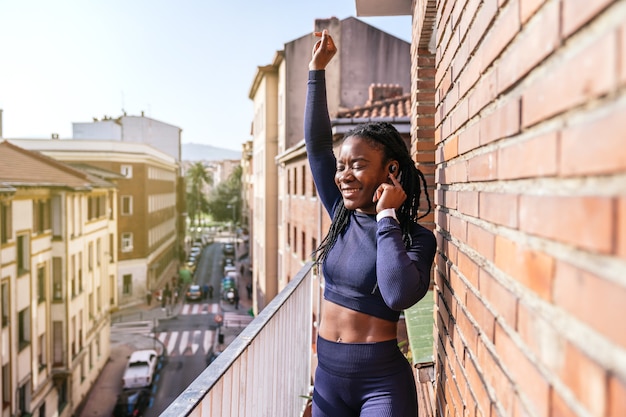 Foto donna afro nera vestita con abiti sportivi che ascolta musica in cuffia molto felice sul balcone perché inizierà ad allenarsi a casa a causa della pandemia di coronavirus covid19