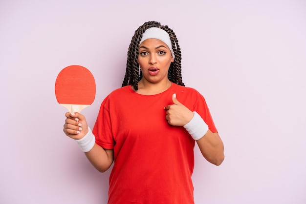 Black afro woman looking shocked and surprised with mouth wide open pointing to self ping pong concept