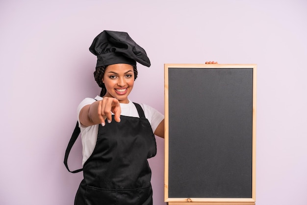 Black afro woman pointing at camera choosing you chef and blackboard concept