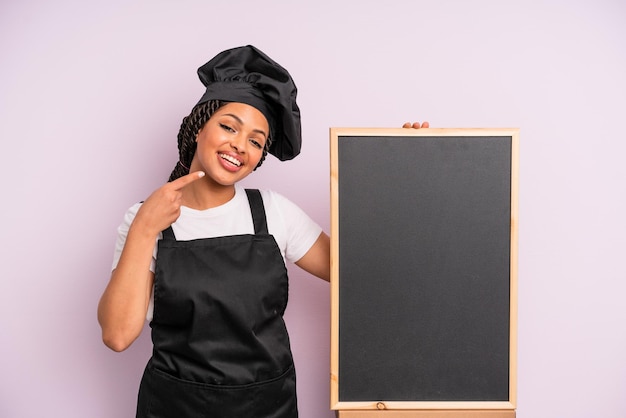 Black afro woman smiling confidently pointing to own broad smile chef and blackboard concept