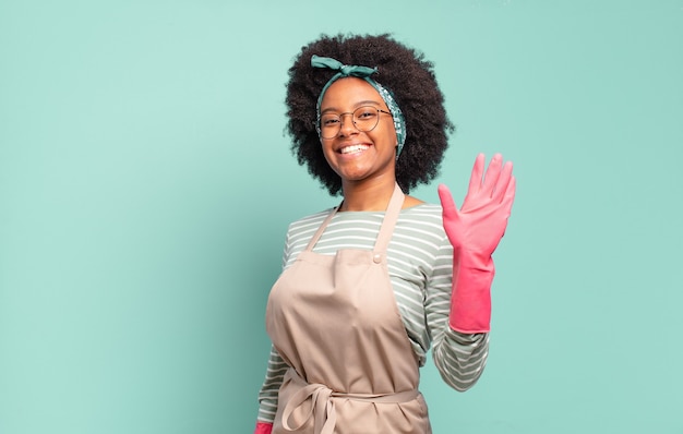 Foto donna afro nera che sorride allegramente e allegramente, agitando la mano, accogliendoti e salutandoti o salutandoti. concetto di pulizia.. concetto di famiglia