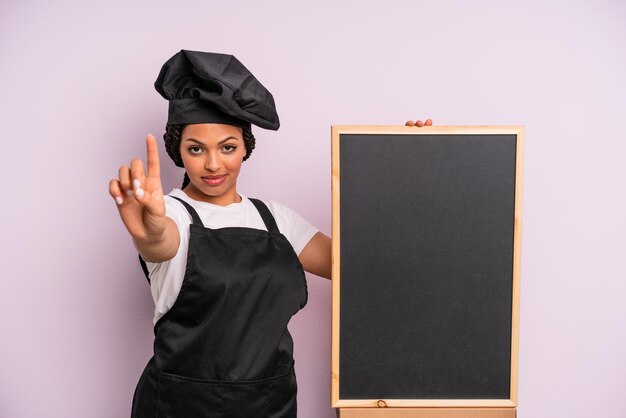 Black afro woman smiling and looking friendly showing number one chef and blackboard concept