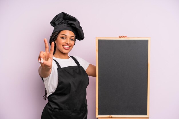 Black afro woman smiling and looking happy gesturing victory or peace chef and blackboard concept