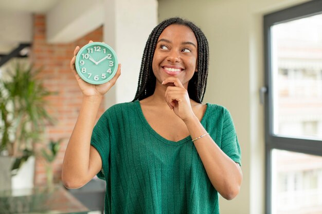 Photo black afro woman smiling with a happy confident expression with hand on chin alarm clock concept