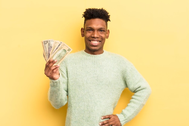 Черный афро молодой человек счастливо улыбается с рукой на бедре и уверенной концепцией долларовых банкнот