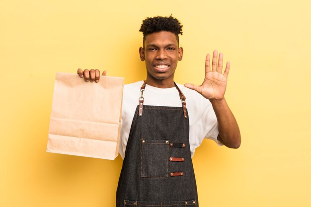 Черный афро молодой человек улыбается и выглядит дружелюбно, показывая номер пять на вынос концепции работника еды