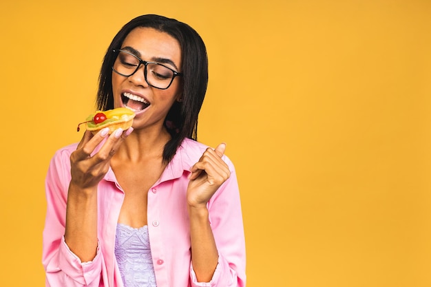 Black american african happy woman eating cake dessert isolated\
over yellow background eating cupcake diet unhealthy food\
concept