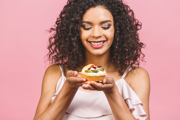 Black american african happy woman with curly afro hair style making a mess eating a huge fancy dessert over pink background. Eating cupcake.