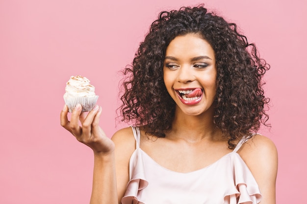 Photo black american african happy woman with curly afro hair style making a mess eating a huge fancy dessert over pink background. eating cupcake.