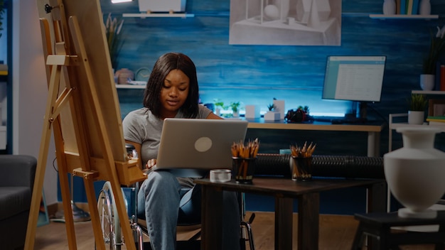 집에 있는 미술실에서 영감을 얻기 위해 노트북 컴퓨터에 타이핑을 하는 핸디캡이 있는 흑인 예술가. 장치와 기술을 사용하여 휠체어를 탄 아프리카계 미국인의 창의적인 여성