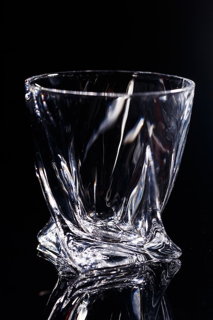 На черном фоне стоит чистый стакан с отражением на стеклянном столе. Грани на стеклянной чашке