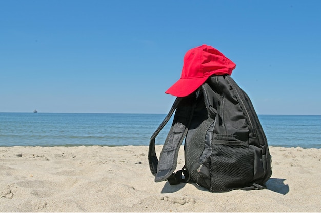 Black backpack and red cap lying on shore of gray sand of calm blue sea