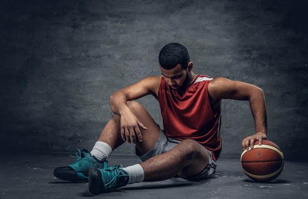 Black basketball player sits on a floor and holds a ball.