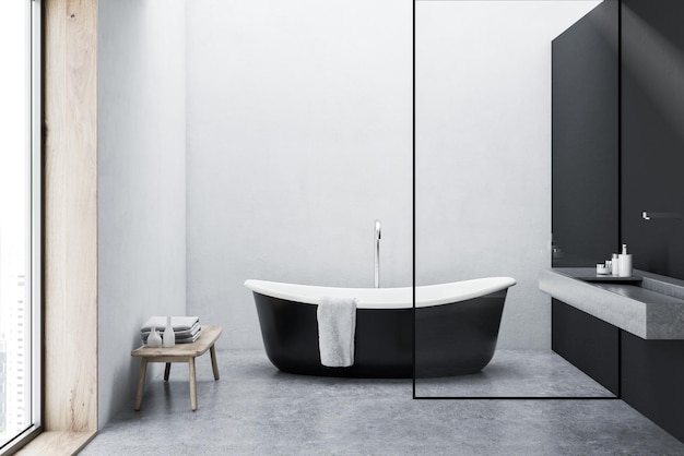 Черная ванна с подвешенным на ней полотенцем, стоящая на бетонном полу минималистической серой и белой ванной комнаты.