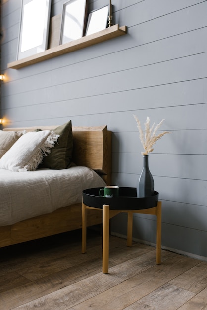 A black bedside table with a gray vase and dried flowers and a green metal mug placed by the bed 