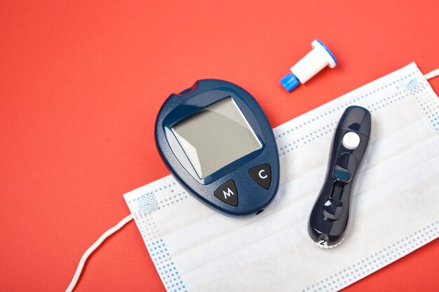 Black blood glucose meter and face mask on a red background, spare needles for the rchka and lancept syringe, protection against viruses of people at risk