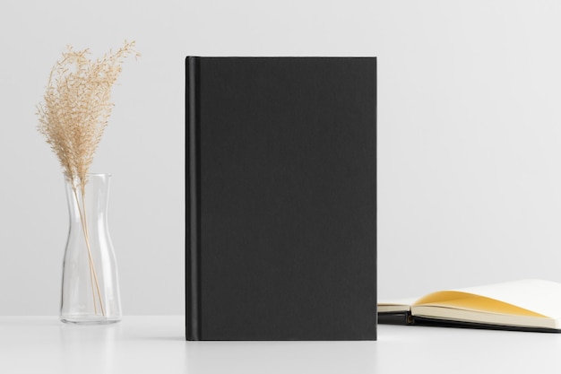 Black book mockup with a dried grass decoration and workspace accessories on a white table