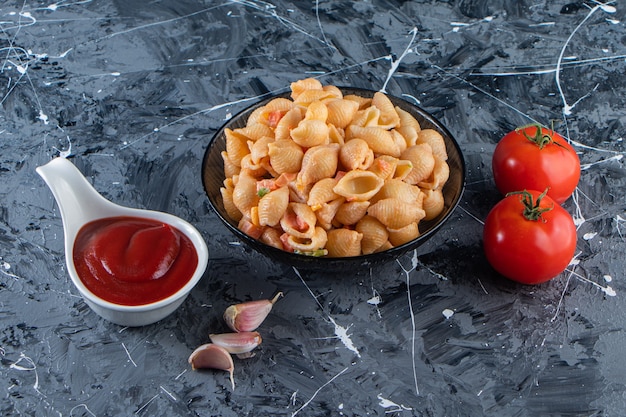 Черная чаша вкусных макаронных изделий из ракушек с кетчупом на мраморной поверхности.