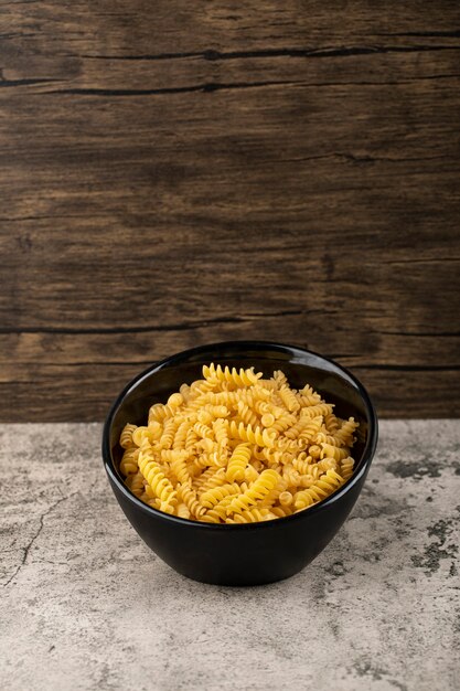 Black bowl of raw fusilli pasta on stone table. 