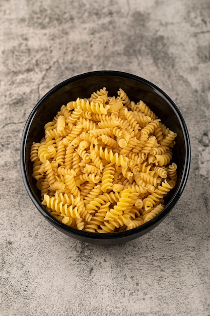 Black bowl of raw fusilli pasta on stone table. 