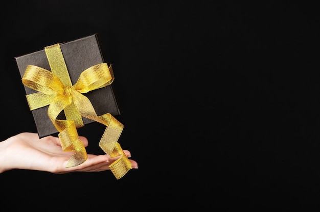 暗い背景に金色のリボンが付いた黒い箱が女性の手の上に浮かんでいます。ブラックフライデーのギフト用ギフトボックス。ホリデーセールのフライングプレゼント。スペースをコピーします。