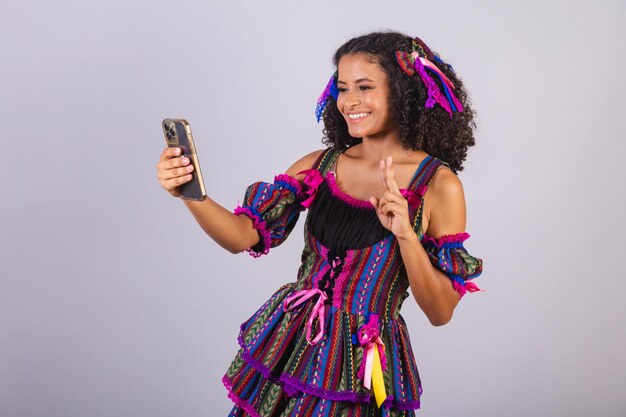 写真 フェスタ ジュニーナの服を着た黒人ブラジル人女性がスマートフォンでチャットする聖ヨハネ アライアルの交わり