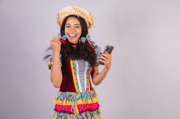 Black Brazilian woman wearing June party clothes fraternization in the name of Sao Joao Arraial with smartphone