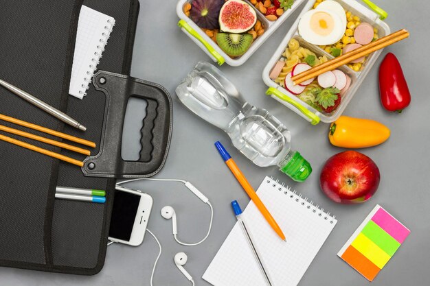 Black briefcase with phone and headphones Bottle of water notebooks with pens lunch box of fruits