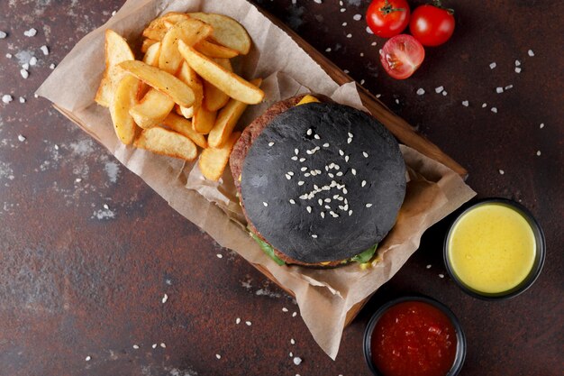 Black bun cheeseburger met sesamzaadjes en gebakken aardappelpartjes in houten kist, dipsauzen set en cherrytomaatjes op rommelige tafel, op oude roestige achtergrond, bovenaanzicht, kopieerruimte. Fastfoodconcept