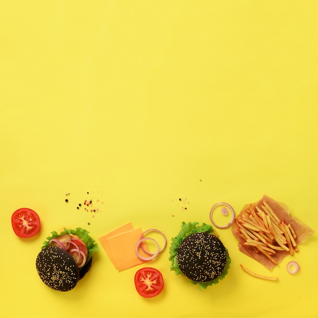 Black burger, french fries potatoes, tomatoes, cheese, onion, cucumber and lettuce on yellow background.Take away meal. Unhealthy diet concept