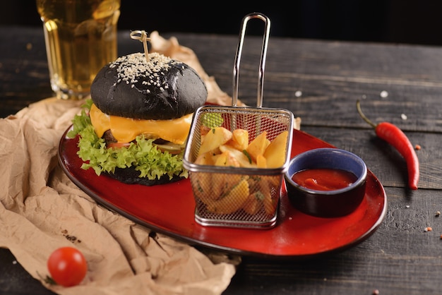 Black burger with fries and sauce. In a red plate on a wooden table