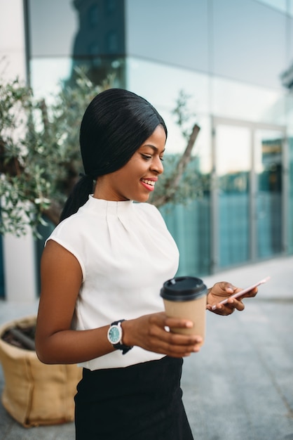 段ボールのコーヒーカップを持つ黒のビジネス女性が事務所ビルに対して携帯電話を使用してください。