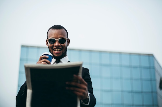 Photo black businessman with coffee in his hand smiles as he reading the newspaper in front of the skyscraper.