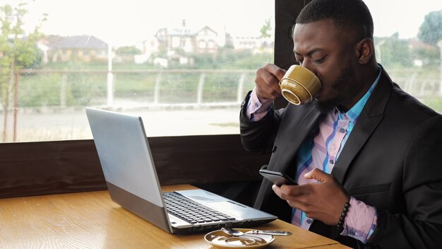 Black businessman works on laptop with smartphone in cafe and drinks coffee.