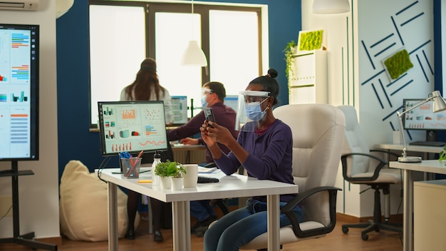 Black businesswoman browsing using smartphone sitting in office room wearing face mask and visor while team doing stategy. Multiethnic coworkers working respecting social distance in financial company