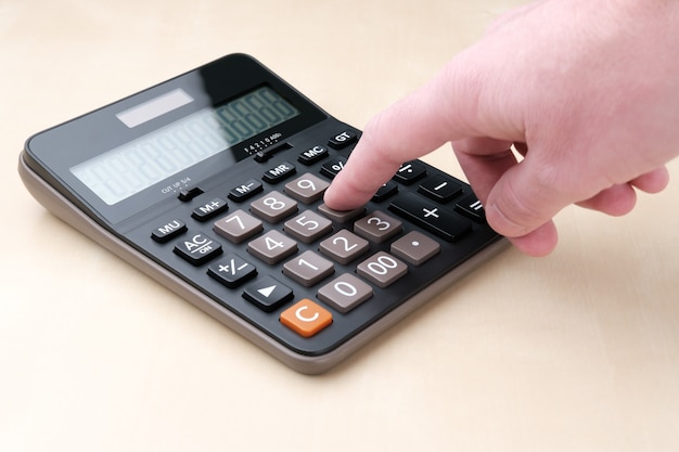 Черный калькулятор с большими кнопками и дисплеем лежит на бежевом столе, и мужчина нажимает пальцем на клавишу с цифрой шесть.