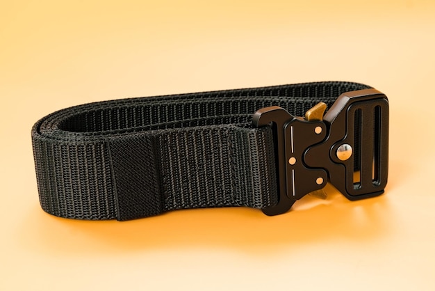A black canvas belt with a black metal male buckle and a black\
belt loop lays on the orange background utility hiking material\
outfit safety soldier steel uniform weapon cloth