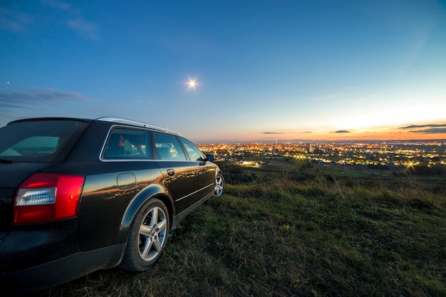 Black car parked at night in green meadows with distant city buildings and bright blue sky.