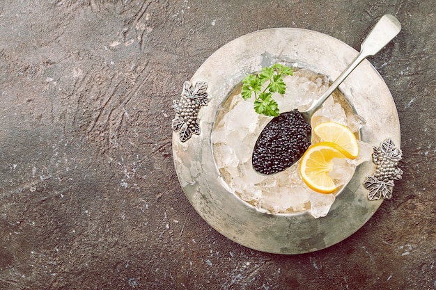 Black caviar in a spoon on ice with lemon close up over dark surface top view