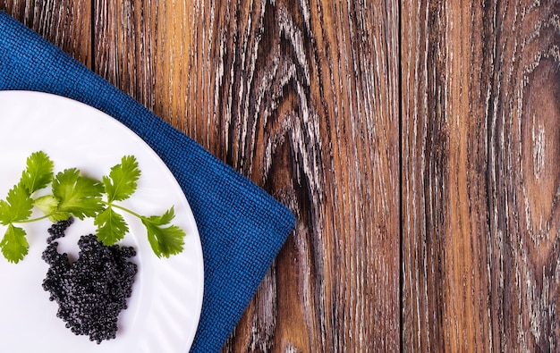 Black caviar on a white plate with herbs on table.