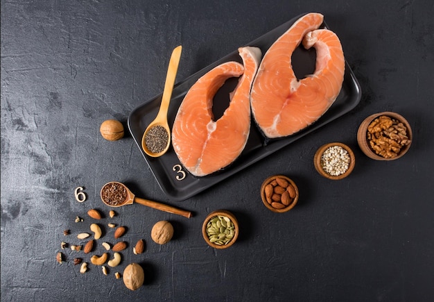 Black ceramic tray with fresh salmon steaks on a black background. wooden spoons and mini bowls filled with nuts, flax seeds, chia, pumpkin.