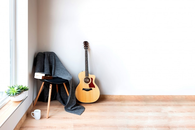 Black chair covered with woolen gray blanket and acoustic guitar in front of an empty white wall. Succulent plants on the window.