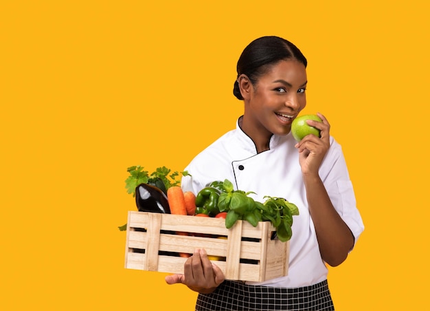 Black chef lady holding crate with vegetables aand fruits and biting apple