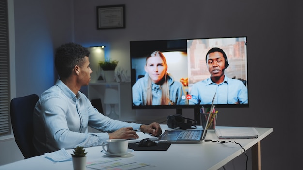 Black chief manager making video call on big monitor with\
colleagues on quarantine