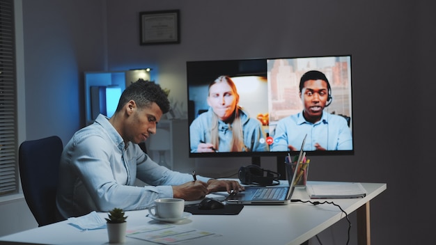 Black chief manager making video call on big monitor with\
colleagues on quarantine