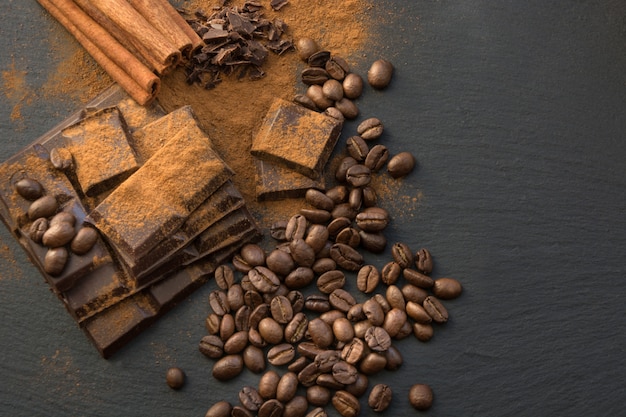 Black chocolate pieces, cinnamon and coffee beans, chocolate slabs spilled from grated chocolate powder on slate dish. Top view.